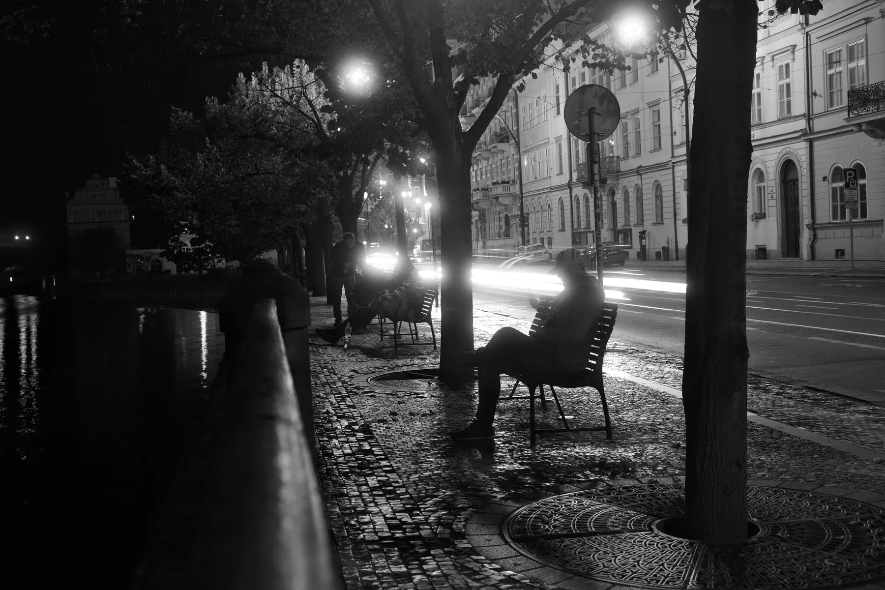 A man on a bench under a streetlamp, wet cobbles, light streaks.