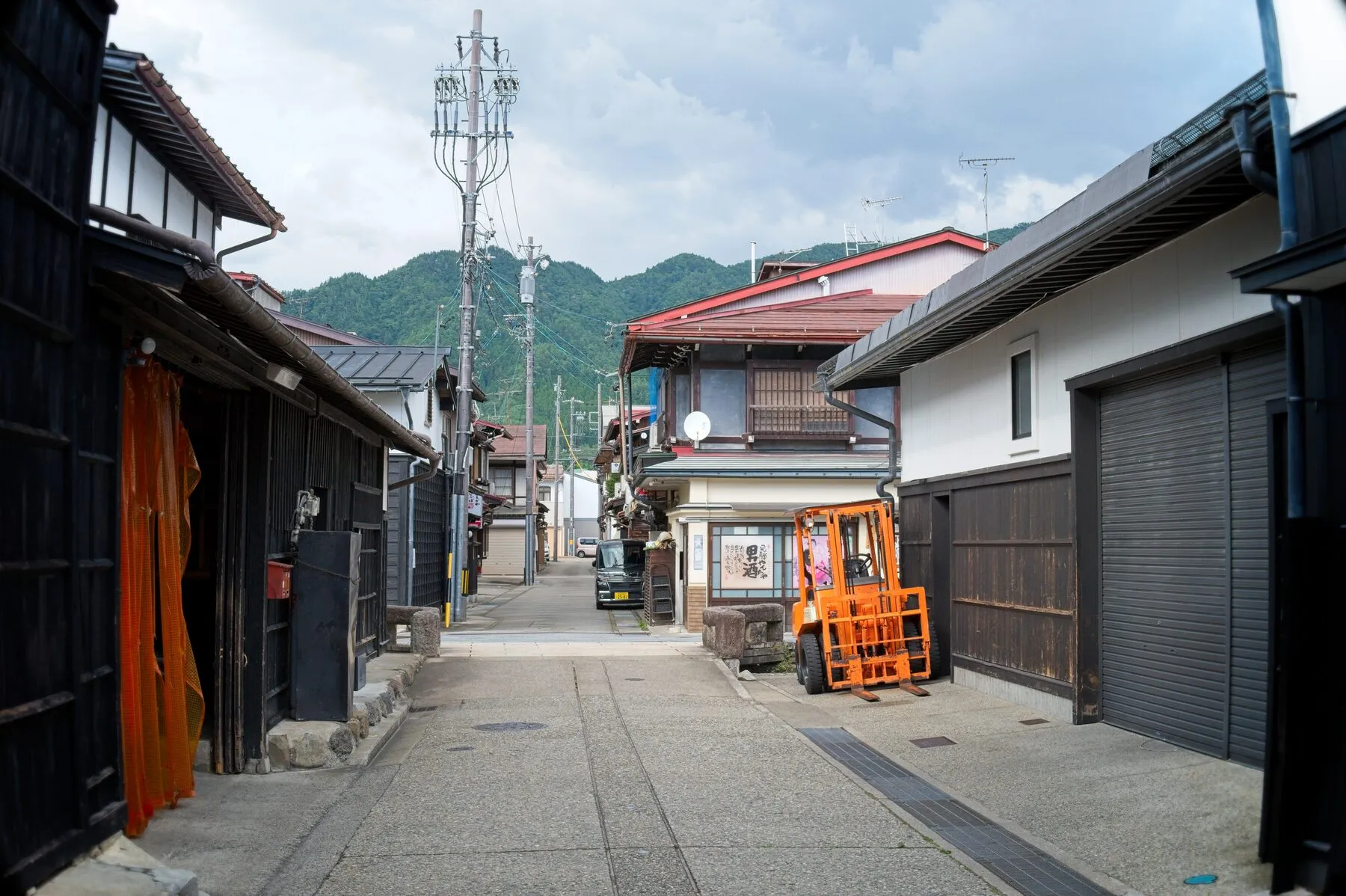 An orange forklift on a sunlit lane of dark-wood houses, hills behind.