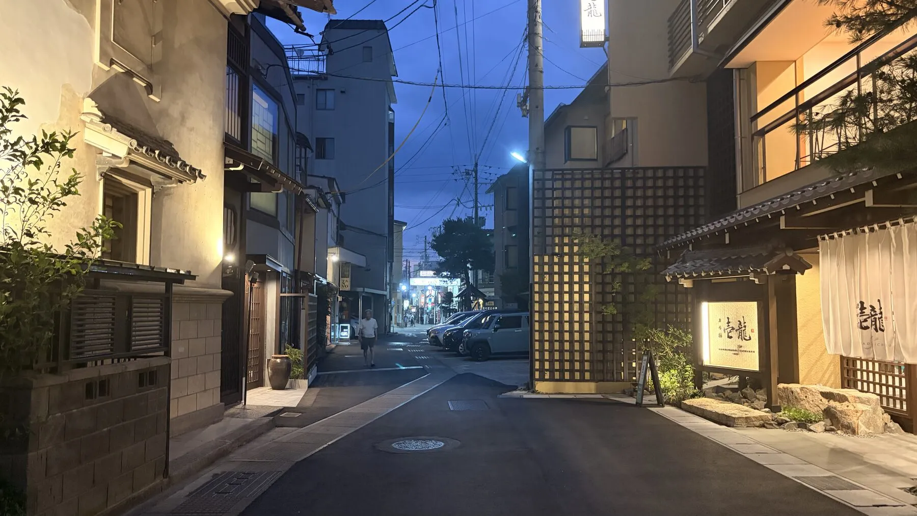 A lantern-lit teahouse lane at dusk, one figure on the wet road.