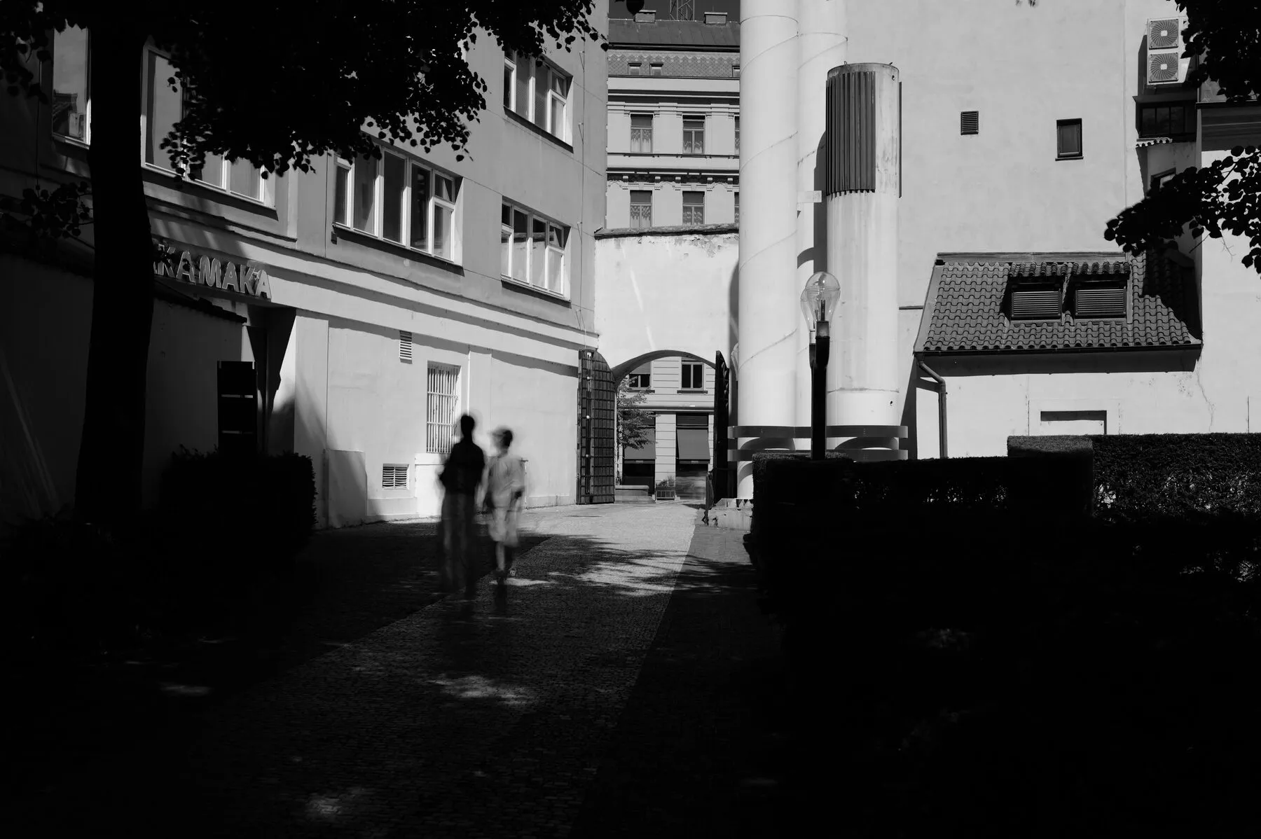 Two blurred figures cross a sunlit plaza, hard shadows.