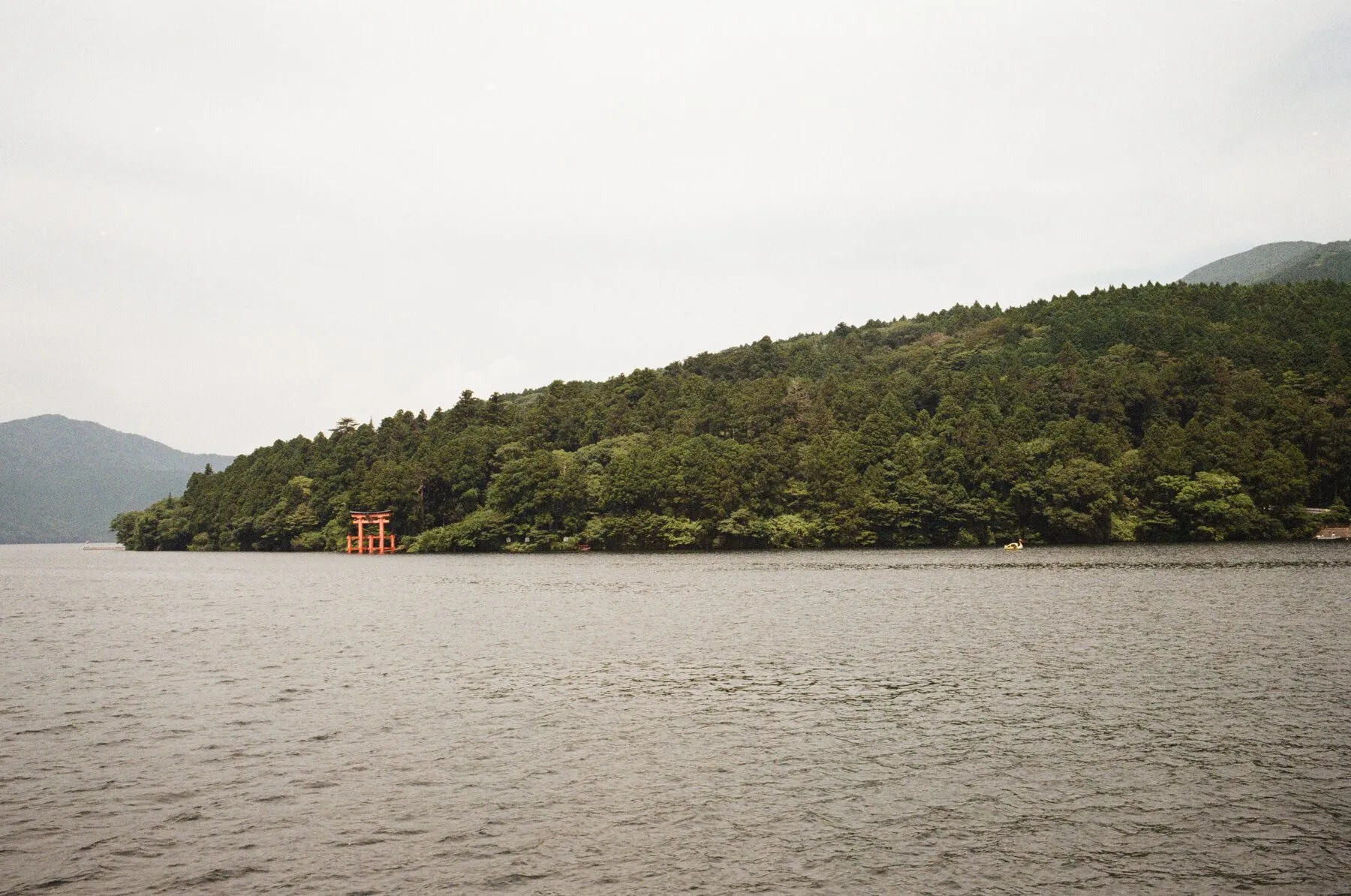 A small red torii on the wooded shore across flat grey water.