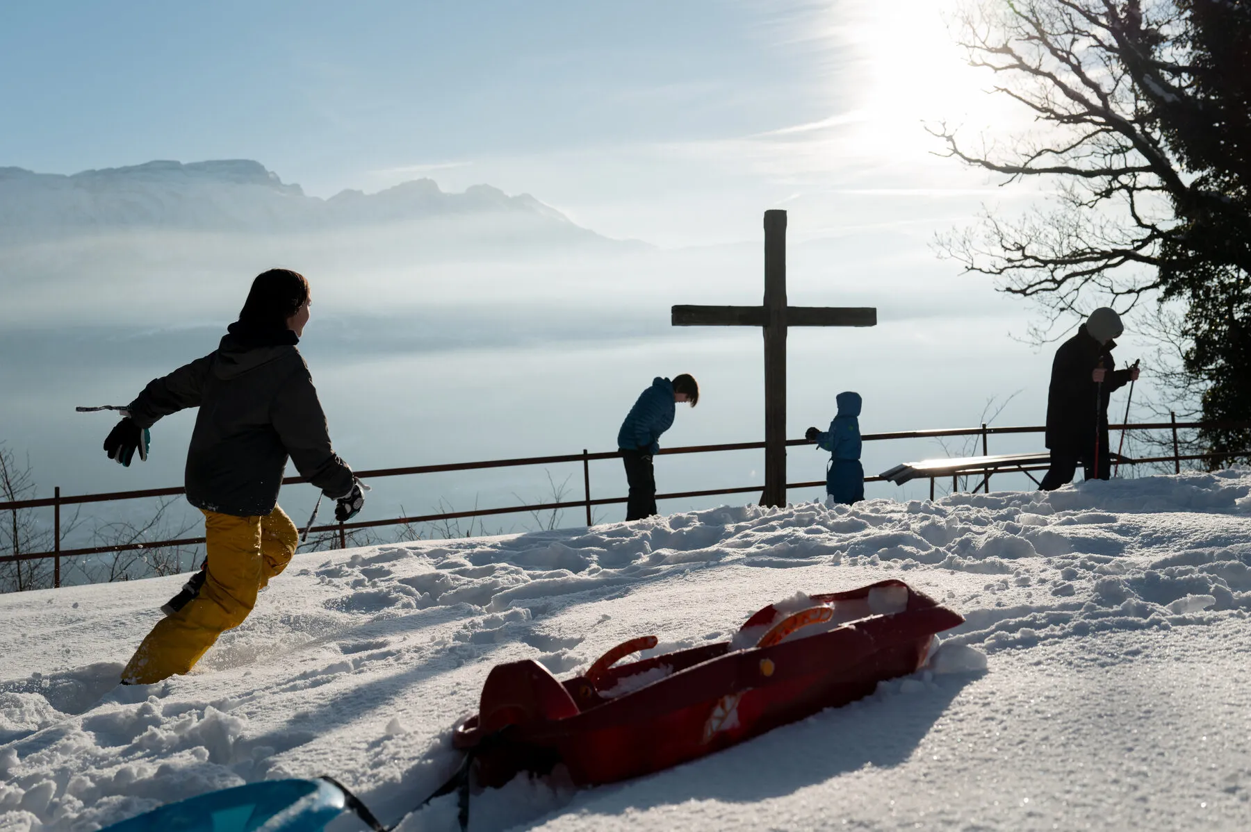 Kids playing by a summit cross, backlit, mist below, sled in the snow.