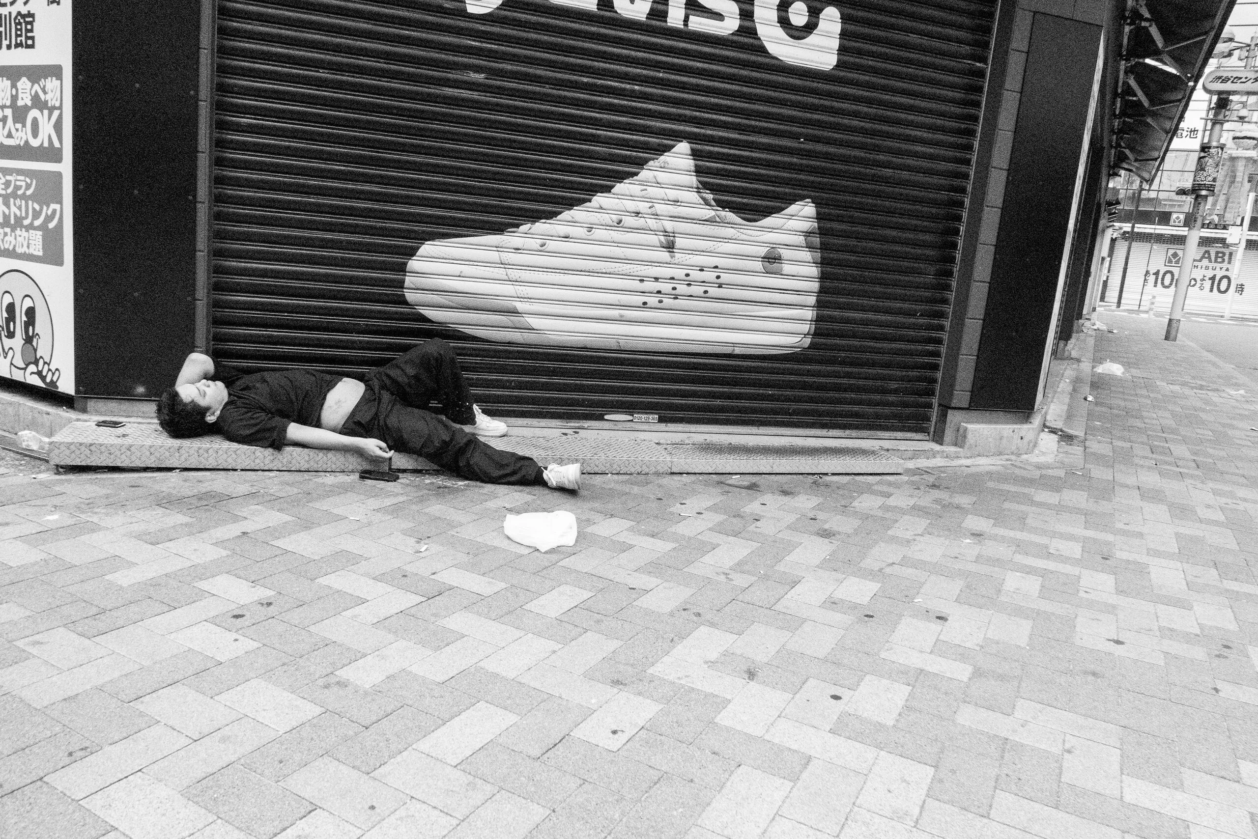 A man asleep on the pavement below a giant sneaker mural.