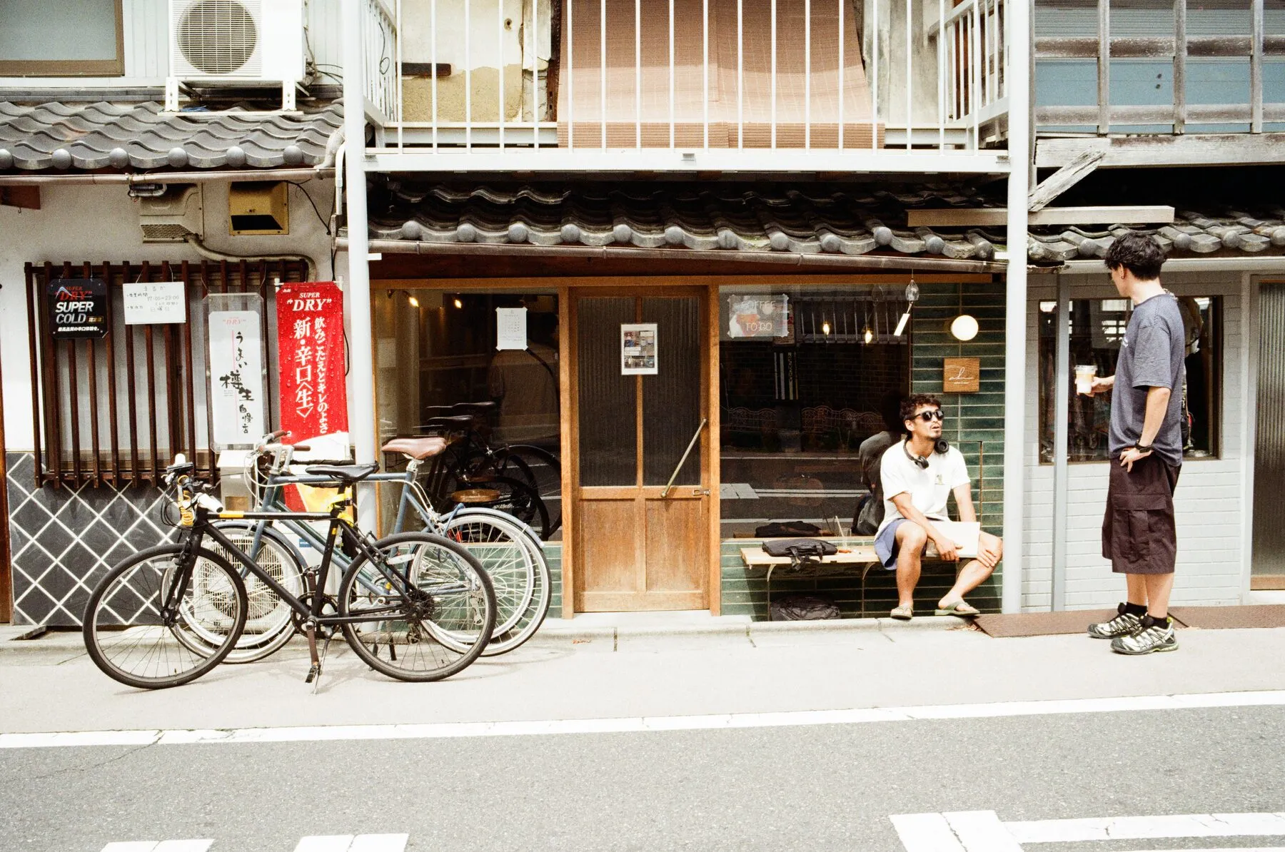 Two young men talk outside a shop, one seated with a coffee, bikes parked.